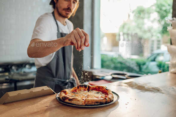 Man worker in a pizza place sprinkling cheese over a baked pizza