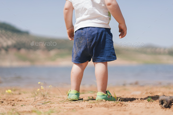 photo of an unrecognizable child with sand-stained clothes, standing by a lake shore