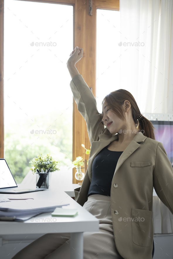Relaxed young female employee sitting at her workplace and stretching her arms.