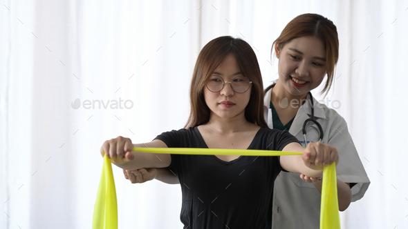 Physiotherapist helping patient exercising arms with yellow elastic rubber at rehabilitation clinic.
