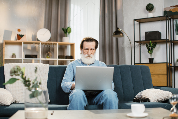 Caucasian pensioner with grey beard in casual wear sitting on comfy couch and working on laptop