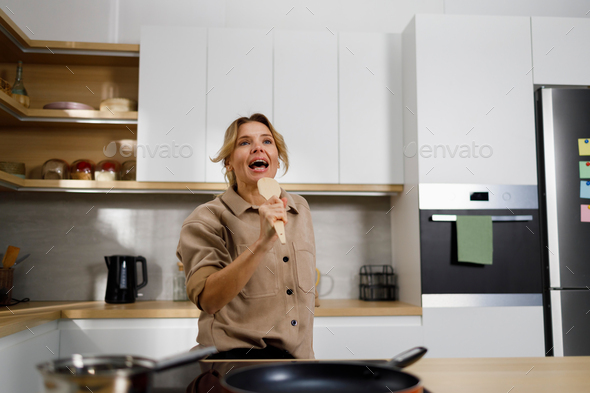 Mature woman cooks food in the kitchen and sings using a wooden spatula as a microphone