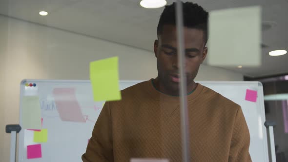 African american businessman standing behind glass wall with sticky notes and pondering alt