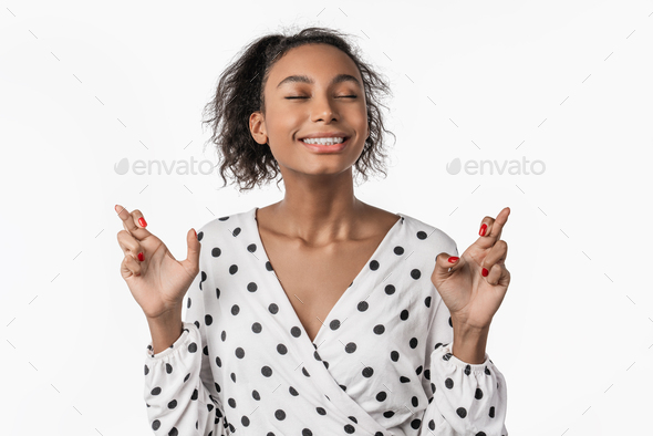 Smiling african woman with closed eyes crossing fingers wishing good luck isolated on studio