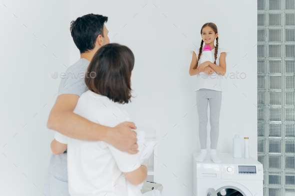 Husband and wife embrace and talk to small girl standing on washer with bottle of detergent.