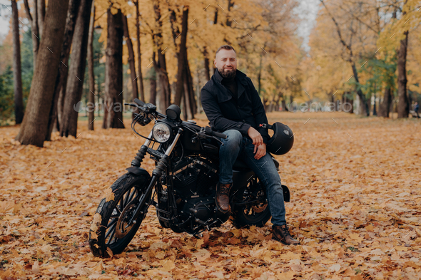 Bearded biker poses on own motorcycle, holds helmet, rides motorbike, poses outdoor in park.