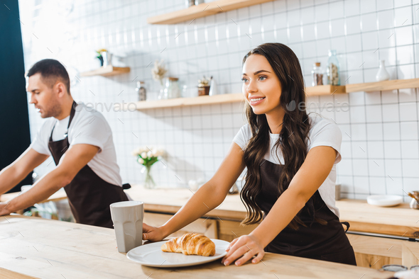 attractive cashier standing behind bar counter with cup, plate and croissant wile barista working in