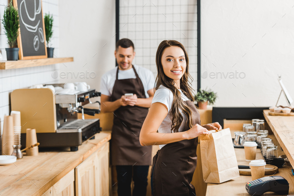 cashier with paper bag smiling and barista with smartphone standing behind bar counter in coffee
