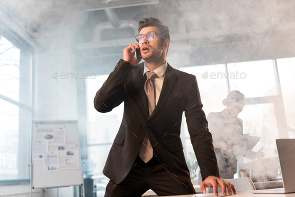 handsome businessman in glasses talking on smartphone in office with smoke near coworker