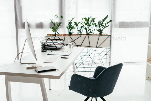 white table with computer, document tray, glass and notebook near office chair and flowerpot with
