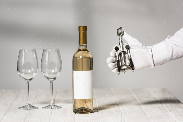 cropped view of waiter in white glove holding corkscrew near table with bottle of wine and wine
