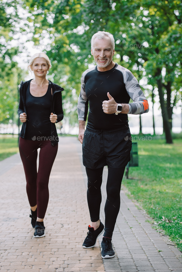 smiling mature sportsman showing thumb up while jogging in park together with attractive sportswoman