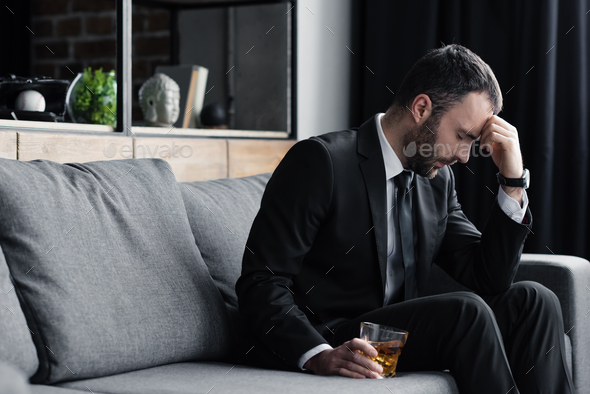 frustrated man in suit sitting on sofa with closed eyes and holding glass of whiskey