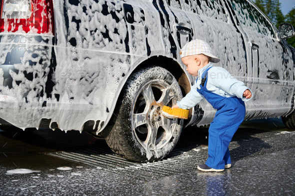 Toddler boy leaning over back wheel of foam-covered car and trying to wash it.