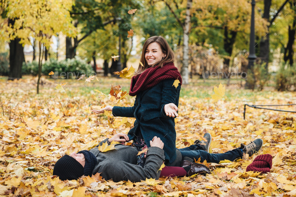 Positive woman wears knitted scarf, sits on boyfriend, throws yellow leaves in air.