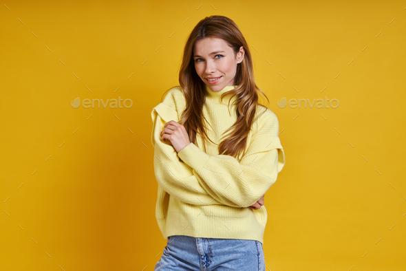 Beautiful young woman in cozy sweater hugging self while standing against yellow background