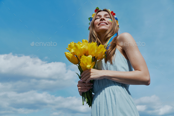 Happy young Ukrainian woman in floral crown holding yellow tulips with blue sky in the background