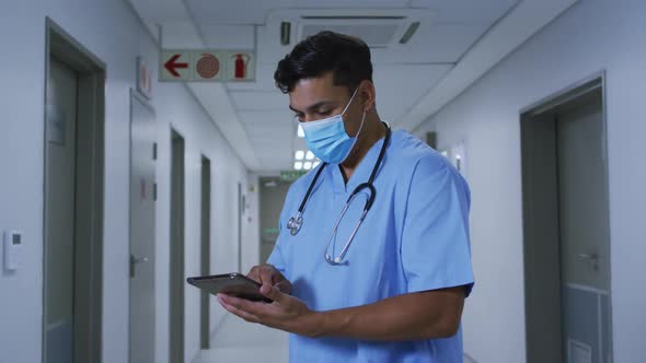 Mixed race male doctor wearing face mask standing in hospital corridor using tablet alt