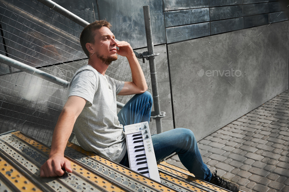 Male with piano keyboard is sitting on stairs outdoors.