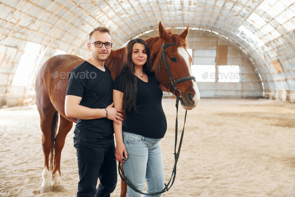 Man with his pregnant wife that is in casual clothes is standing with the horse on a stable