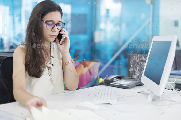 Young woman with cellphone watching computer screen at work