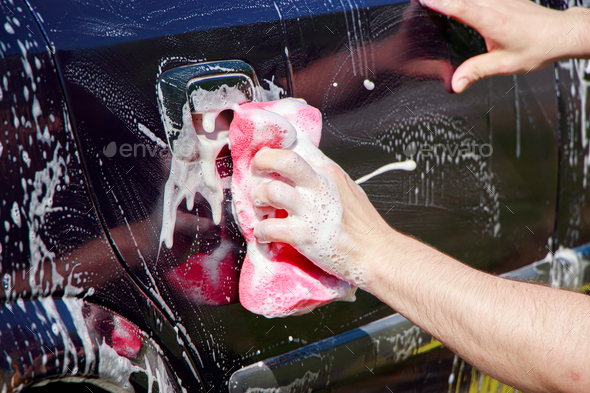 Man is washing car outside. Hand cleaning car. Male hand holding pink sponge with foam.