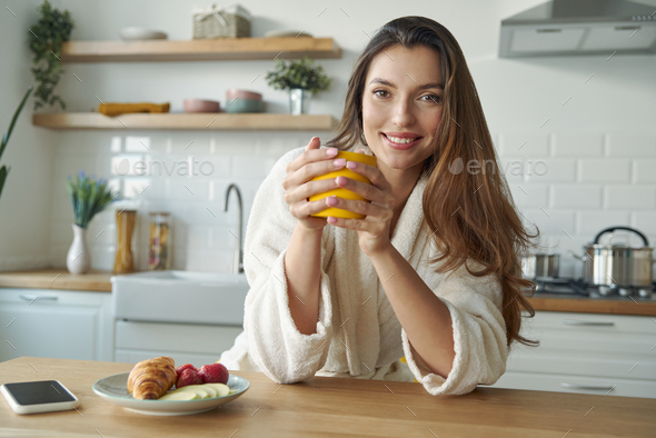Cheerful young woman in bathrobe enjoying breakfast while sitting at the kitchen counter at home