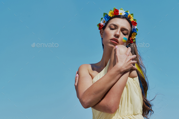Sad Ukrainian woman in floral crown hugging self with sky in the background