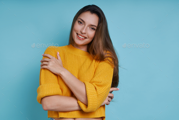 Beautiful young woman hugging self and smiling while standing against blue background