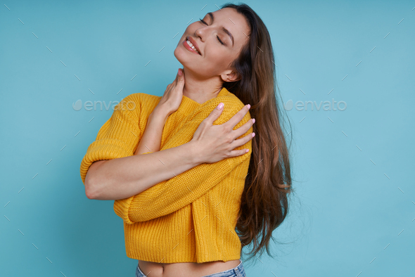 Attractive young woman hugging self and smiling while standing against blue background