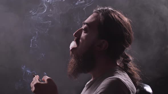 A Man with a Beard and Long Hair Smokes a Shoal in a Dark Room a Lot of Smoke Around Him alt
