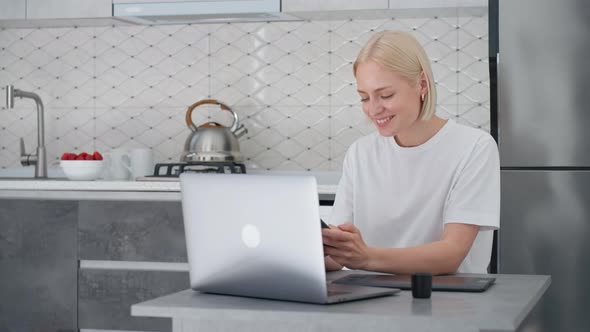 Young Blonde Woman Looks at Smartphone and Smiles While Working From Home