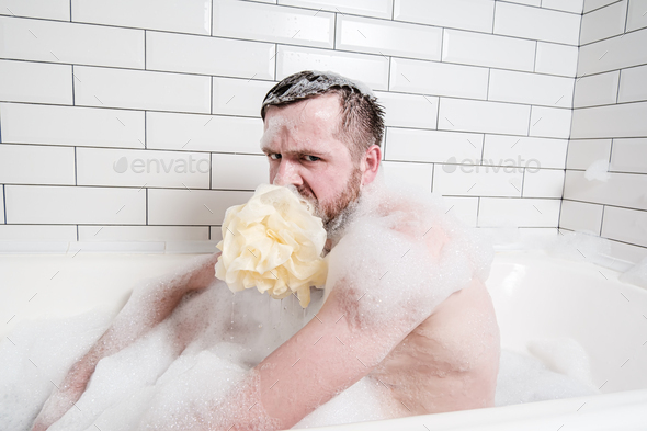 weird man with soapy sponge puff in mouth, sits in bathtub with lush foam and looks angrily