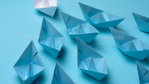 Group of blue paper boats follow white against a light blue background