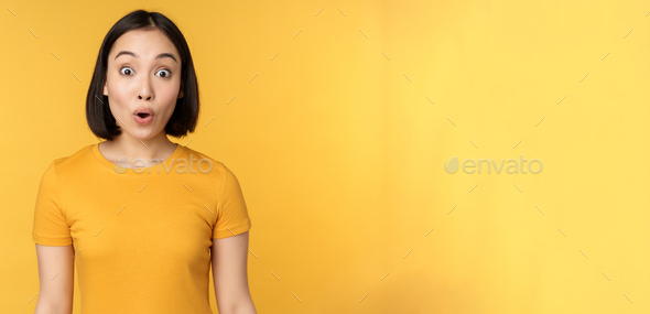 Image of asian girl looking surprised, reacting amazed, raising eyebrows impressed, standing over