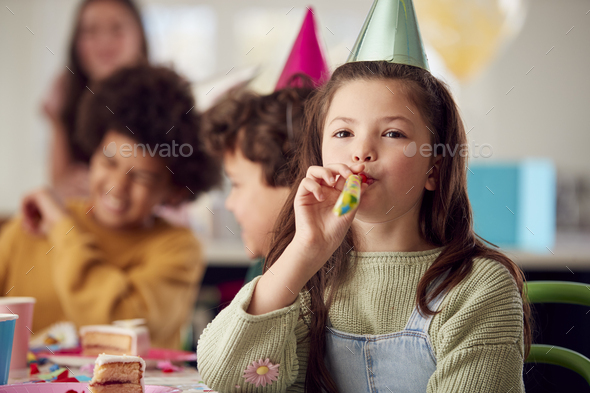 Girl With Birthday Cake And Party Blower At Party With Parents And Friends At Home