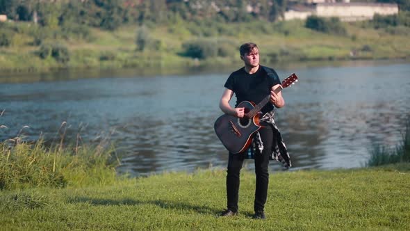 A Male Guitarist Plays an Acoustic Guitar Against the Backdrop of a River