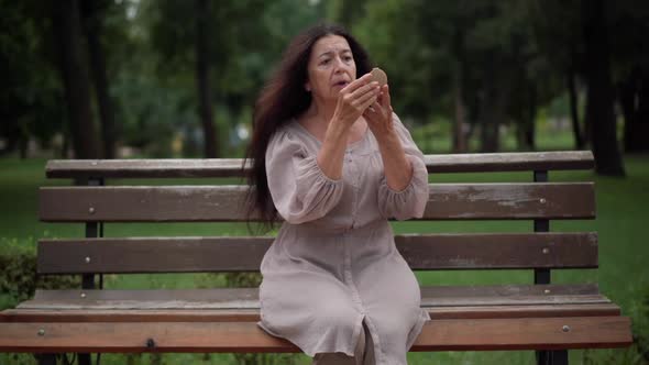 Front View Wrinkled Senior Caucasian Woman Looking in Hand Mirror Sitting on Bench in Park Sighing alt