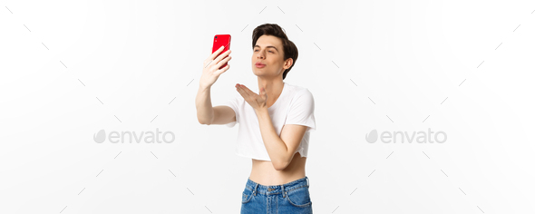 Beautiful gay man in crop top sending air kiss at phone camera, taking selfie or video chat on