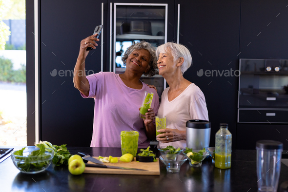 African american senior woman taking selfie with asian friend while holding smoothie in kitchen