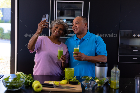 African american senior woman taking selfie with caucasian man while having smoothie in kitchen