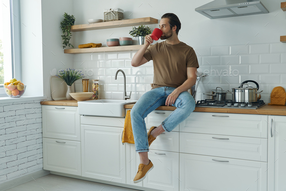 Handsome young man enjoying coffee while sitting on the kitchen counter at home