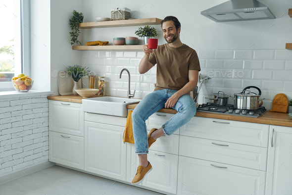 Cheerful man enjoying morning coffee while sitting on the kitchen counter at home