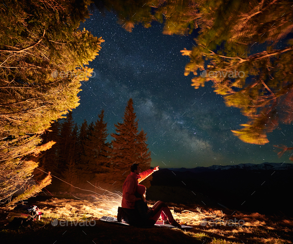 Male traveler showing starry sky to girlfriend in mountains near fire.