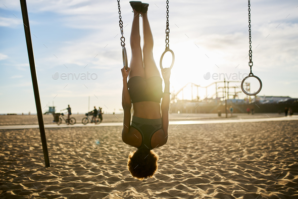 fit african american woman flipping upside down on gymnastic rings at exercise park