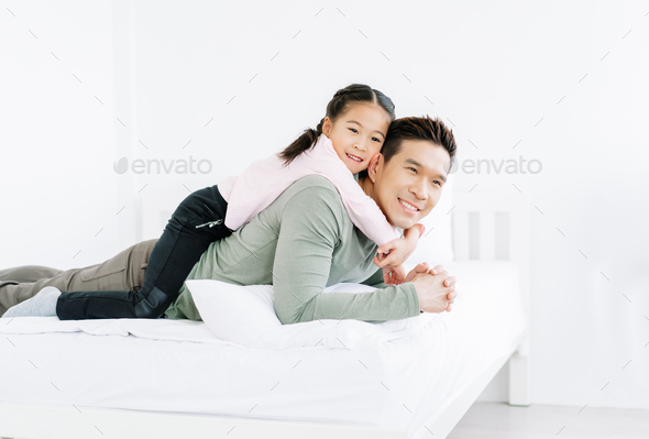Lovely Asian daughter smiling and lying on her handsome father\'s back on white bed.