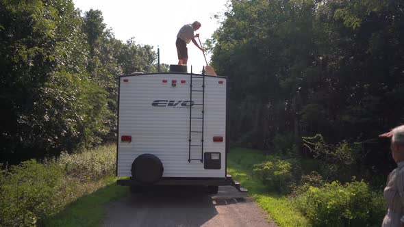 Elderly man sweeping the top of his RV with a broom after boondocking. alt