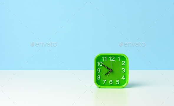 Square green clock with white numbers on a white table, ten minutes to eight in the morning