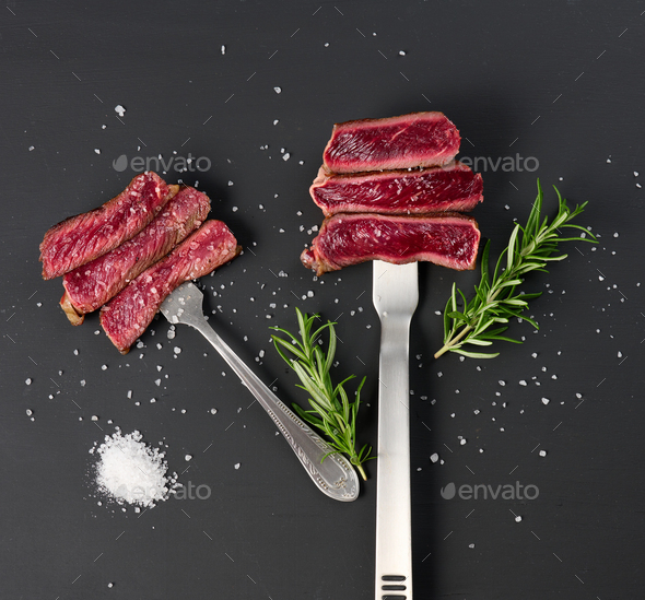 Fried pieces of beef on an iron fork on a black background. The degree of readiness is rare