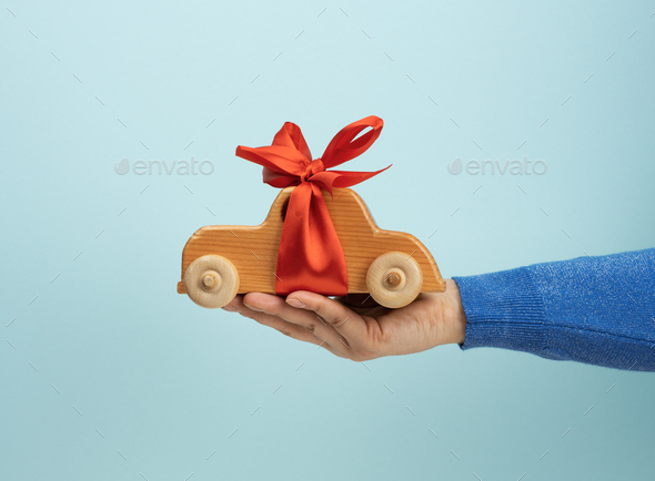 Female hand holding wooden toy car with red ribbon on blue background, auto insurance concept, loan